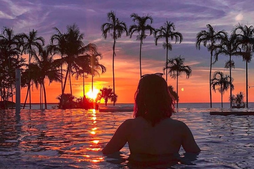 Silhouette of a woman standing chest-deep in water looking at a palm-tree lined sunset from a beachside infinity pool.