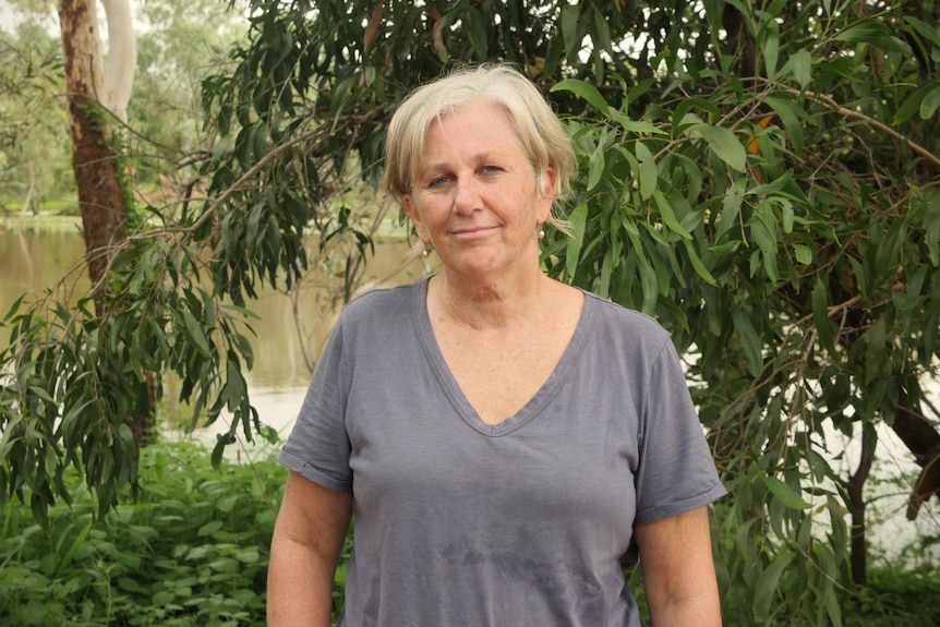 A woman in a grey v-neck shirt stands in front of a green bush and gum tree by the Katherine River