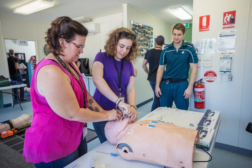 Two women practicing how to check breathing on a dummy as a paramedic watches on.