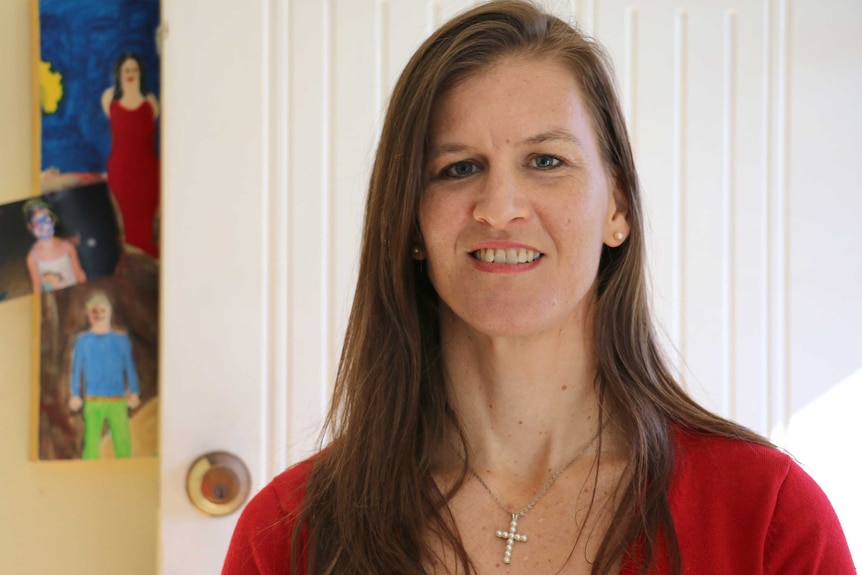 A head and shoulders shot of a smiling woman with long brown hair posing for a photo indoors in front of children's drawings.