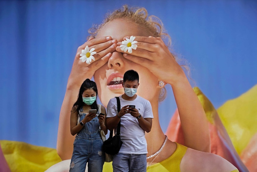 A couple wearing face masks look at their mobile phones in front of a big poster of a girl.