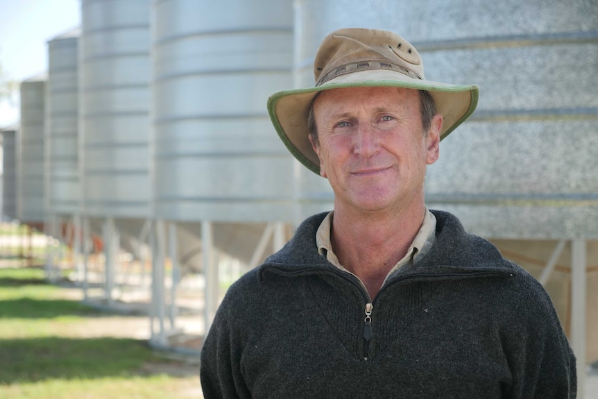 A middle-aged man in a farmer's hat stands near some grain silos looking at the camera