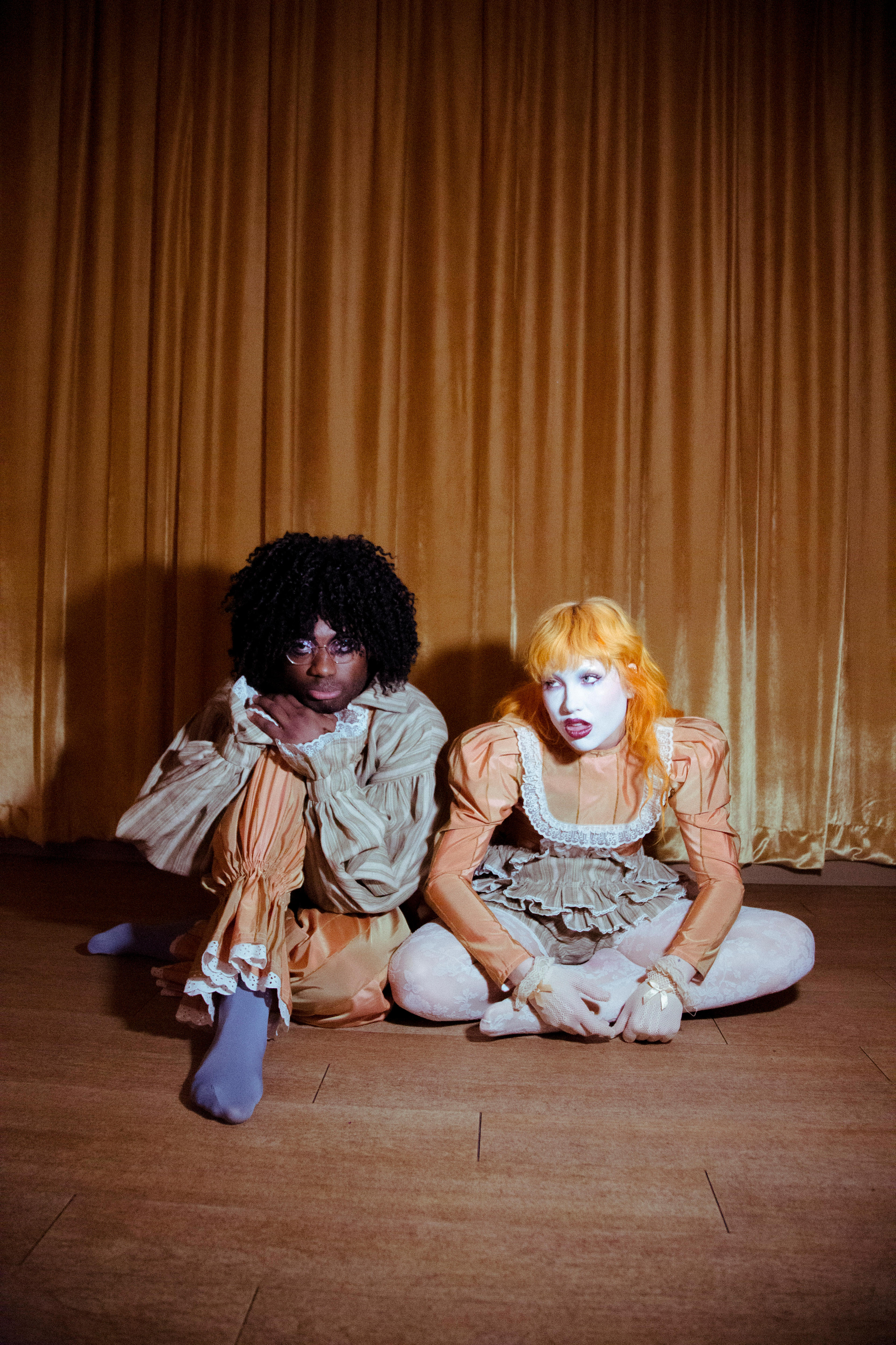 A Black man with a black afro and a white woman with apricot coloured hair sit together on a wooden floor in clown costumes