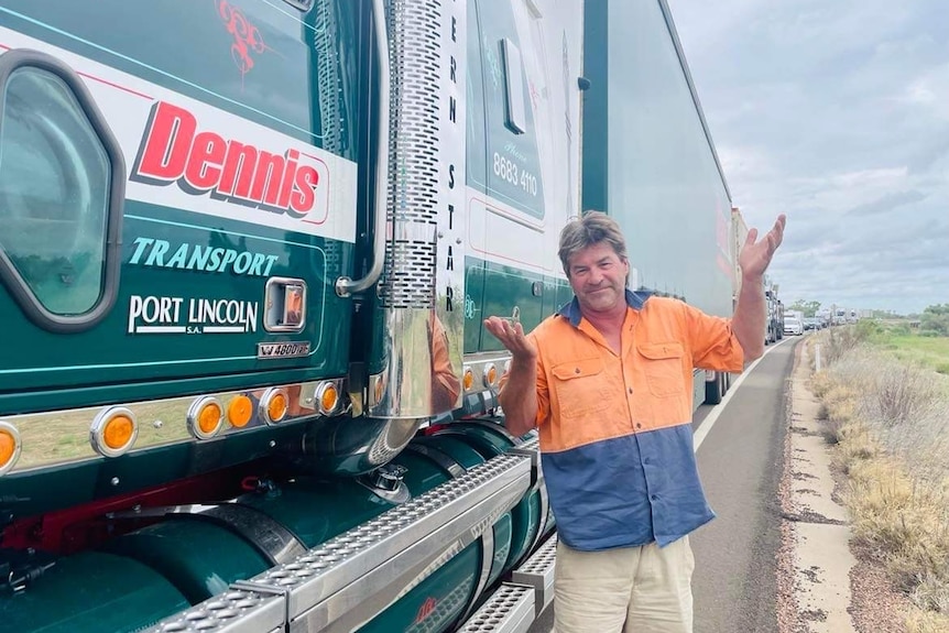 A truck driver wearing orange shrugs his shoulder beside his truck, with congested traffic in the background.