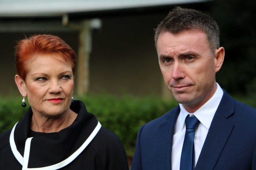 Pauline Hanson looks at a grim looking James Ashby at a press conference. The background is out of focus.