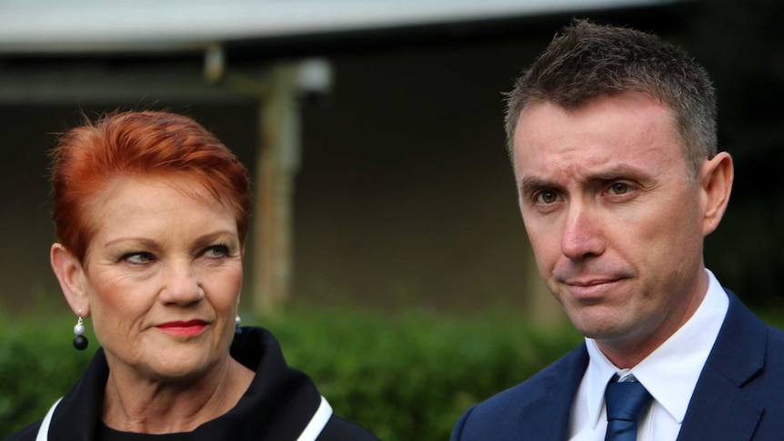 Pauline Hanson looks at a grim looking James Ashby at a press conference. The background is out of focus.