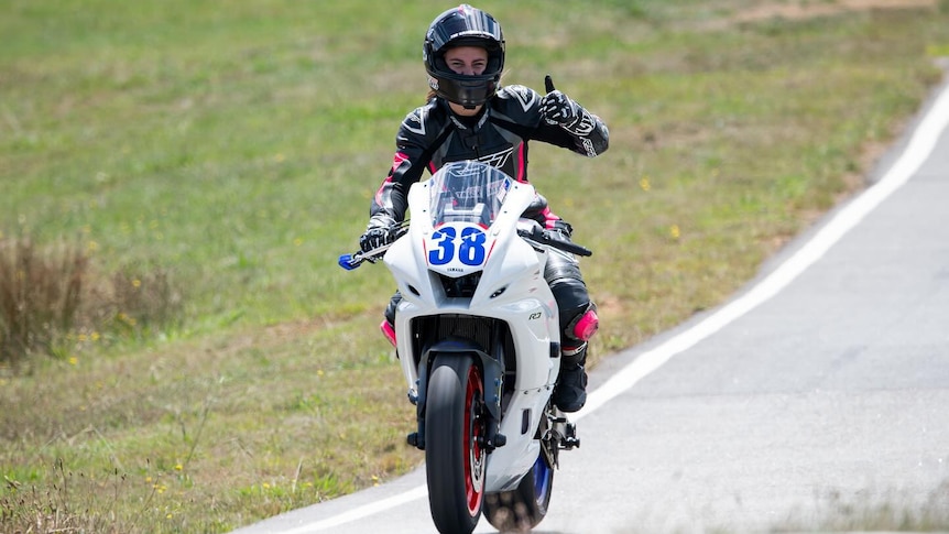 A woman in racing gear sits on a motorbike, grinning through her helmet and giving a big thumbs up 