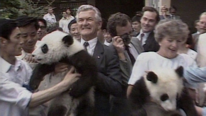 Bob Hawke and Hazel Hawke hold two panda cubs as people watch and take photos.
