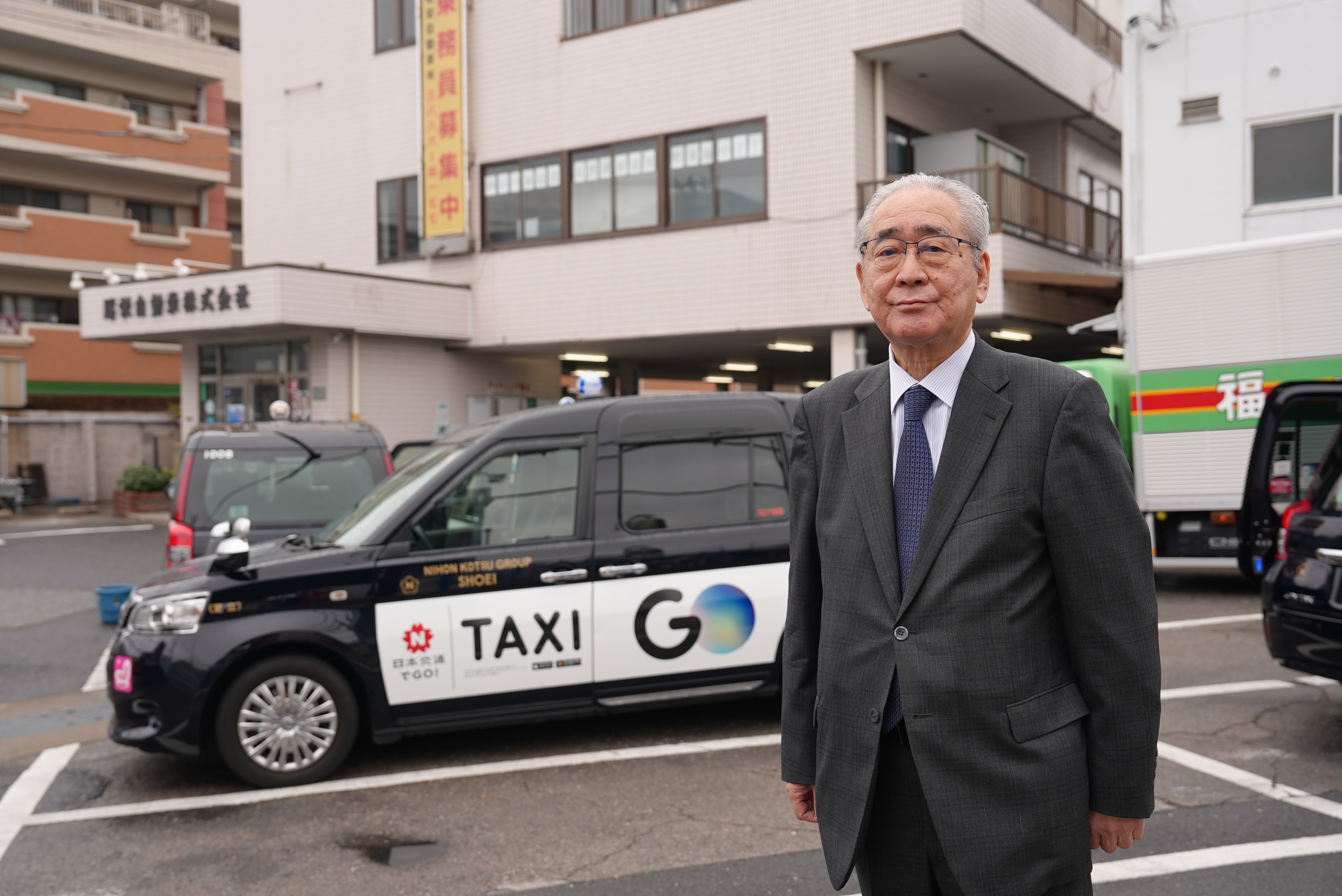 A close up of a Japanese man wearing a dark suit standing infront of a taxi. 