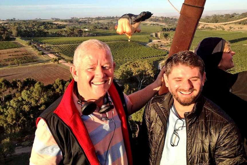 Two men smile at the camera from inside a hot air balloon floating over vineyards