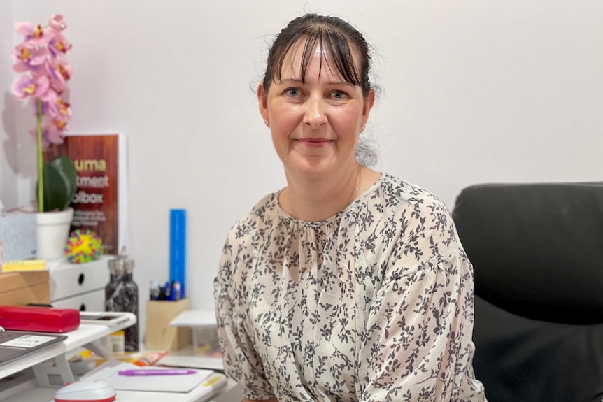 A woman in a black and white floral dress sits at a desk and smiles at the camera