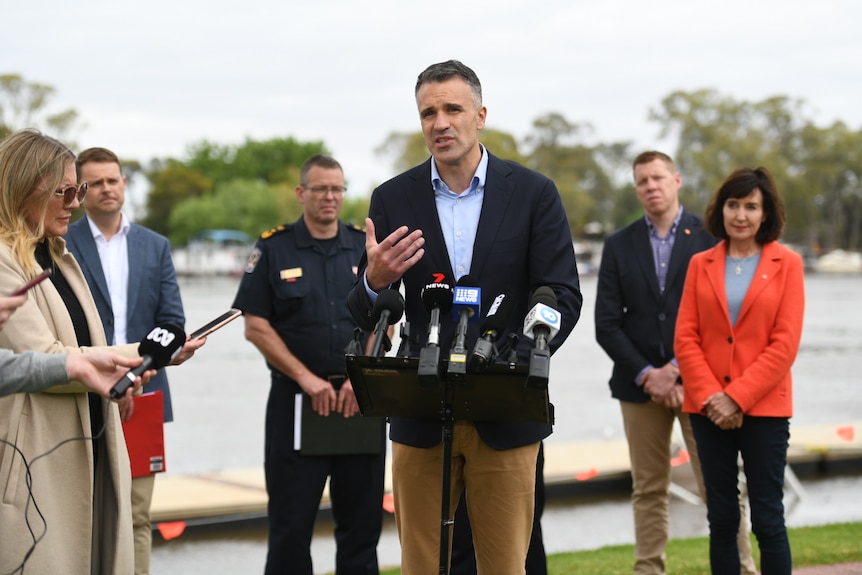 A man wearing a blazer and chinos speaks to microphones while other people stand behind him