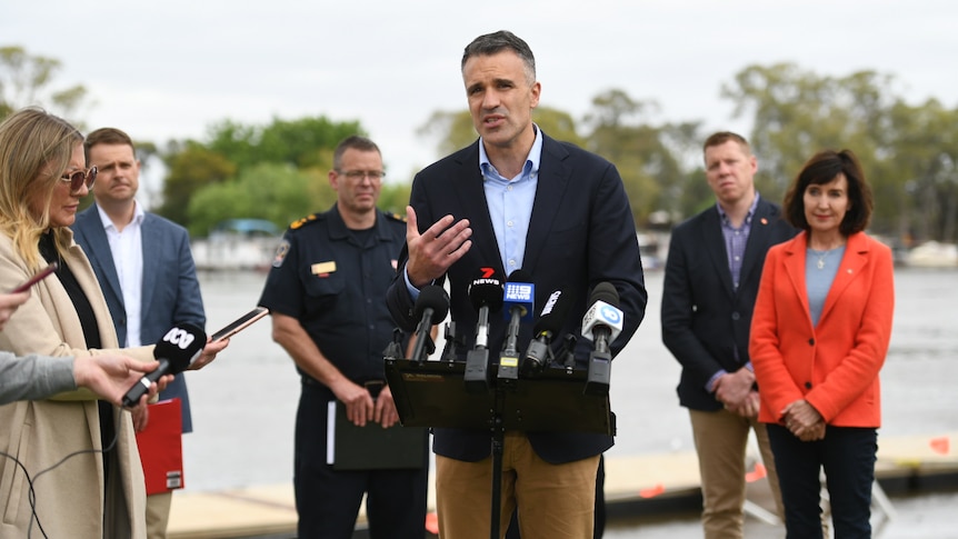 A man wearing a blazer and chinos speaks to microphones while other people stand behind him