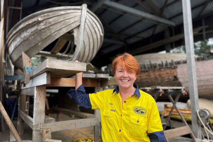 A woman in a bright yellow work shirt stands in front of the hull of an old timber boat, being restored.