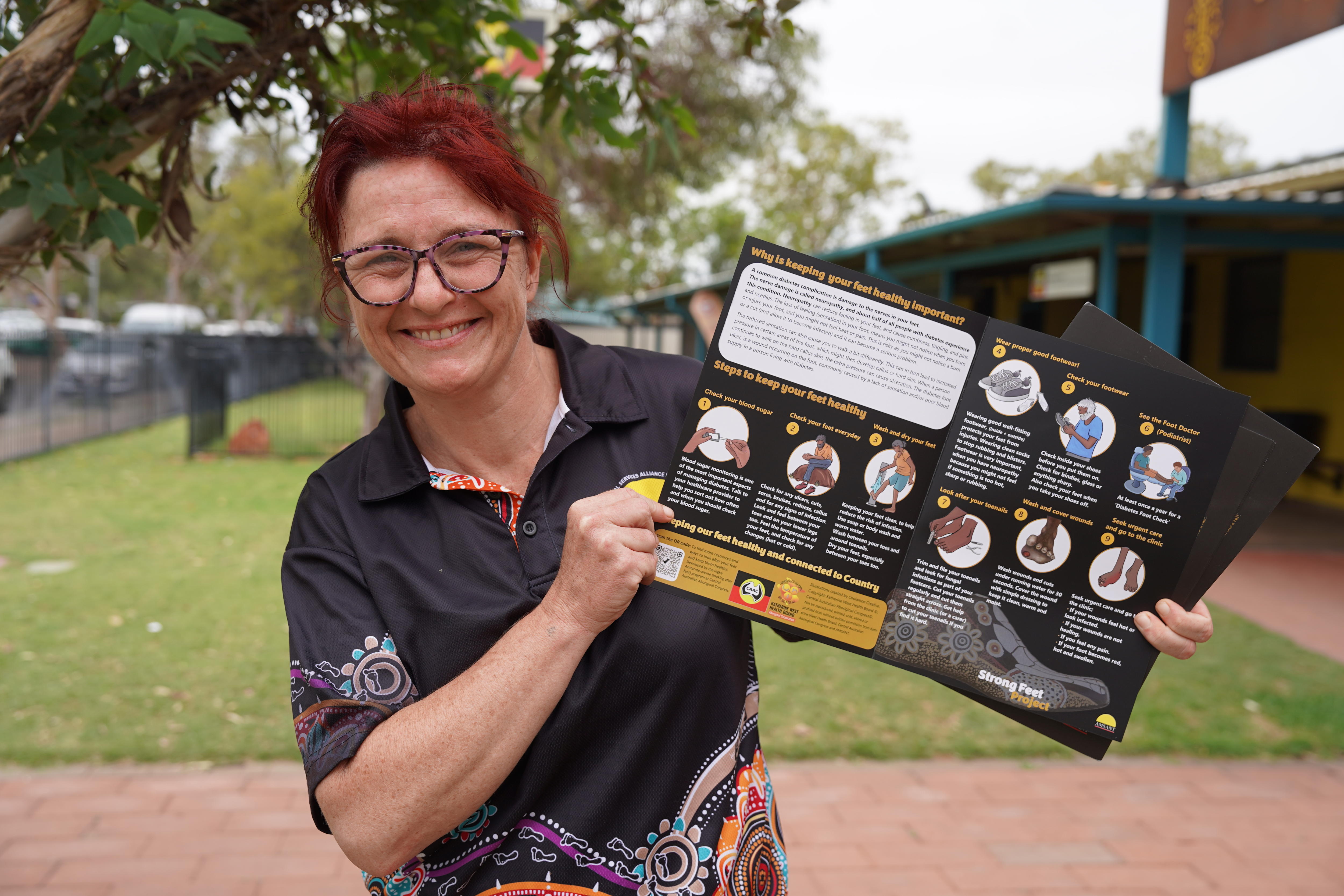 Uma mulher, vestindo uma camisa pólo preta e cabelo ruivo preso para trás, sorri enquanto segura um folheto aberto no gramado da frente do prédio.
