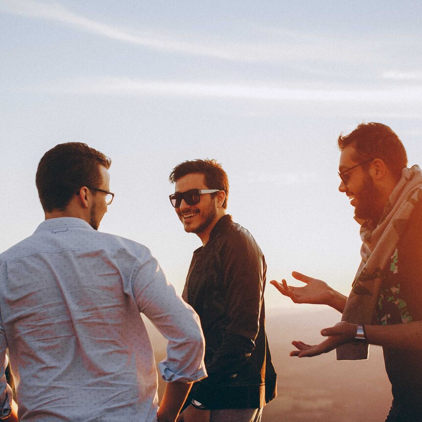 Three young men stand laughing together against a scenic vista.