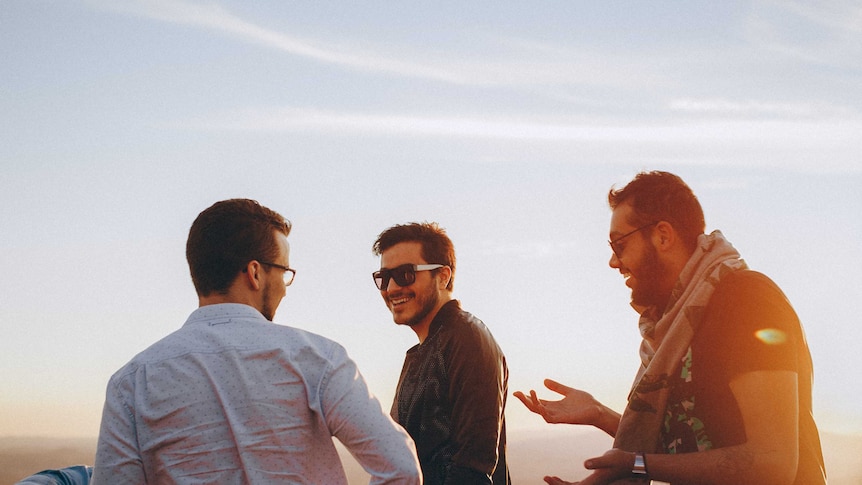 Three young men stand laughing together against a scenic vista.