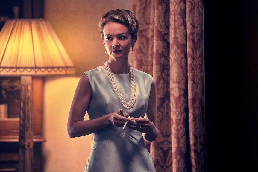 A photo of a white woman with blonde hair in a 50s-style light-blue dress and pearls holding a cigarette