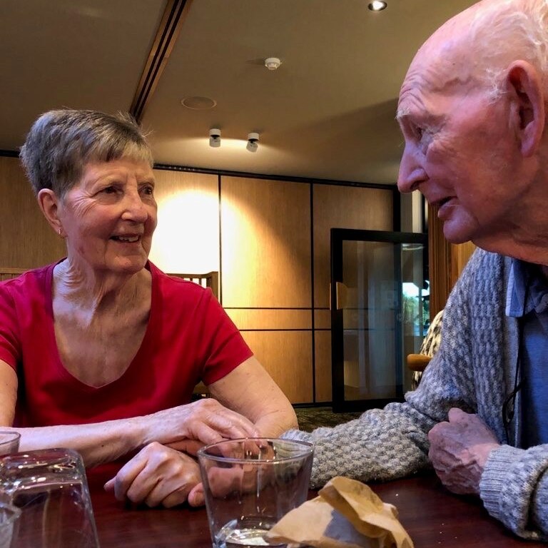 Two elderly people hold hands and talk at a table.
