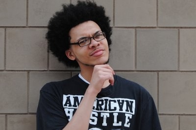 a man holds his hand under his chin while wearing glasses and a black shirt and standing up against a brick wall