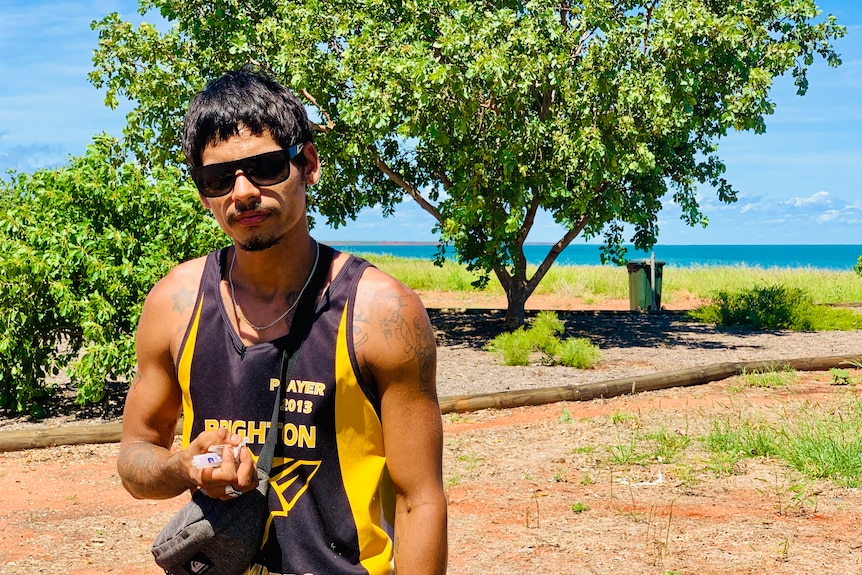 Man man with dark hair and sunglasses stands in front of blue ocean, holding a handful of needles.