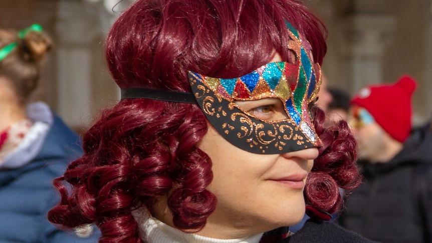 A woman with red hair wears a Venetian mask, in front of a crowd of others also wearing masks.