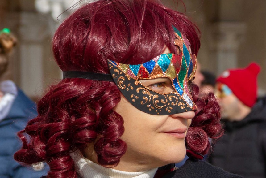 A woman with red hair wears a Venetian mask, in front of a crowd of others also wearing masks.