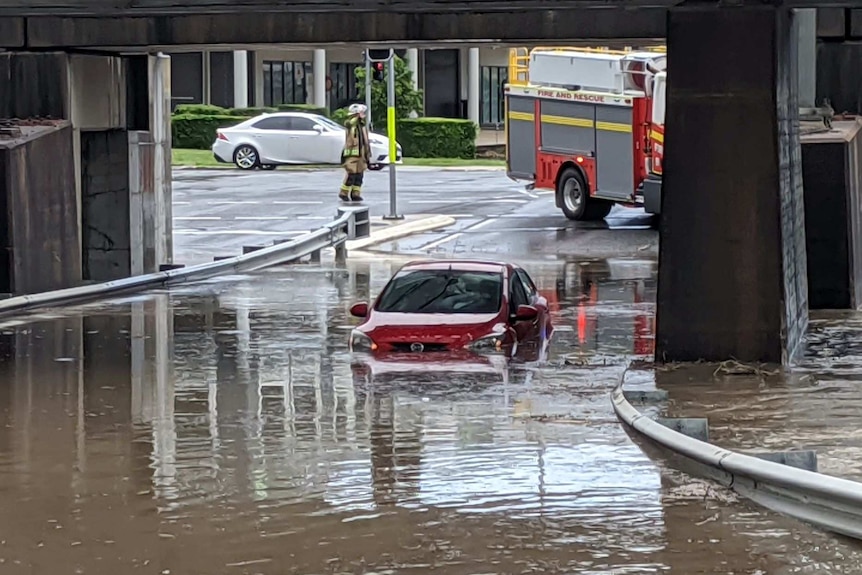 A red car is almost fully submerged in water under a bridge and a fire truck is behind it