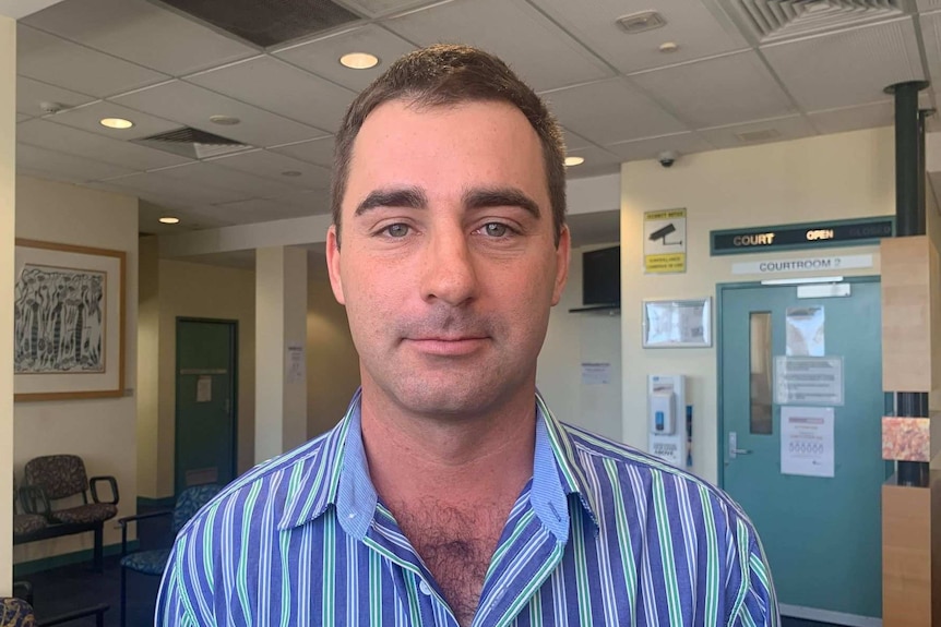 A man in a striped shirt stands, staring directly at the camera. He is in a court house.