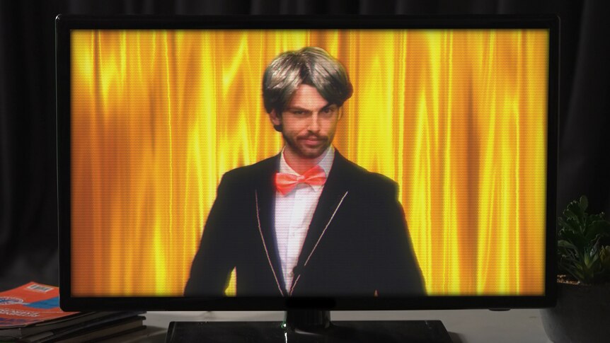 Josh, dressed as a game show host, smirks at the camera from within a TV.