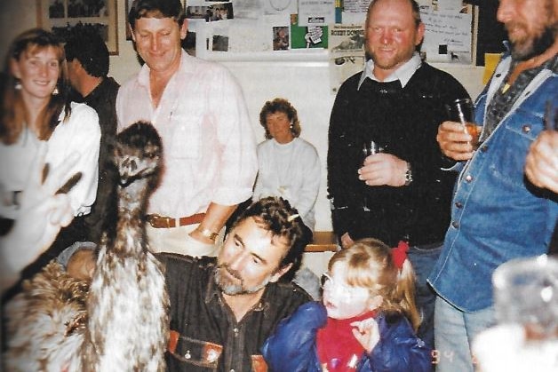 People in a pub, man and girl seated in foreground, man has arm on emu's back, emu on left faces camera