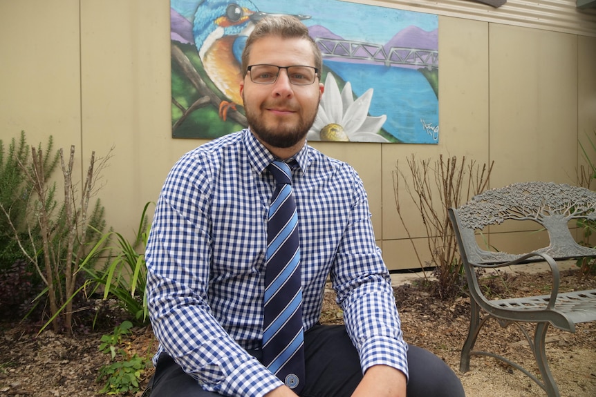 Man in checked blue shirt and tie in sits in front of a painting on the wall behind in courtyard 