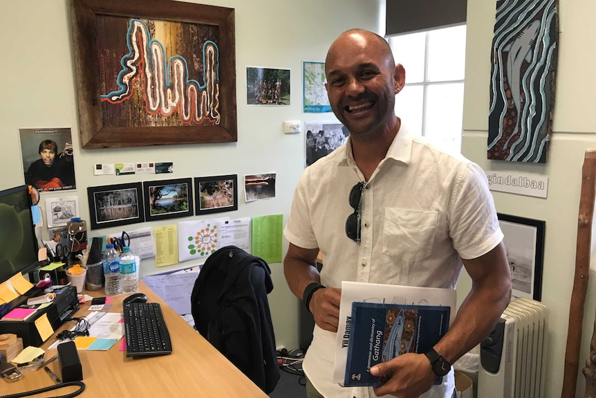 A teacher smiles from behind his desk with Aboriginal art on the walls behind him.