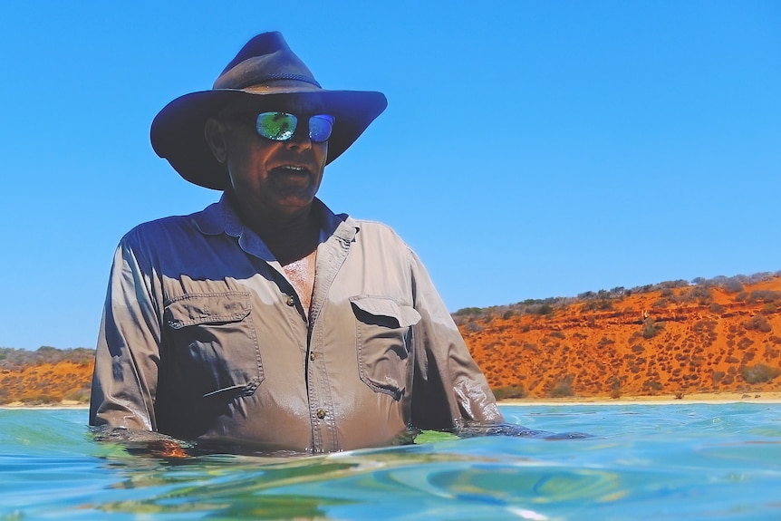 A man in the ocean wearing a large hat and sunglasses. 
