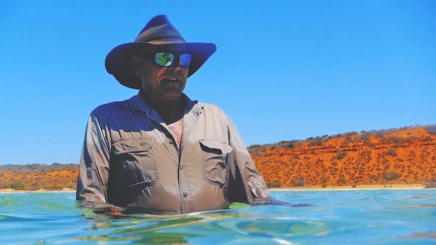 A man in the ocean wearing a large hat and sunglasses. 