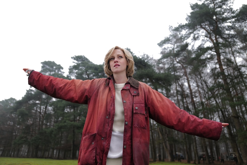 A distressed-looking 30-something woman with a blonde bob, wearing an oversized red parka, stands arms outstretched in a field