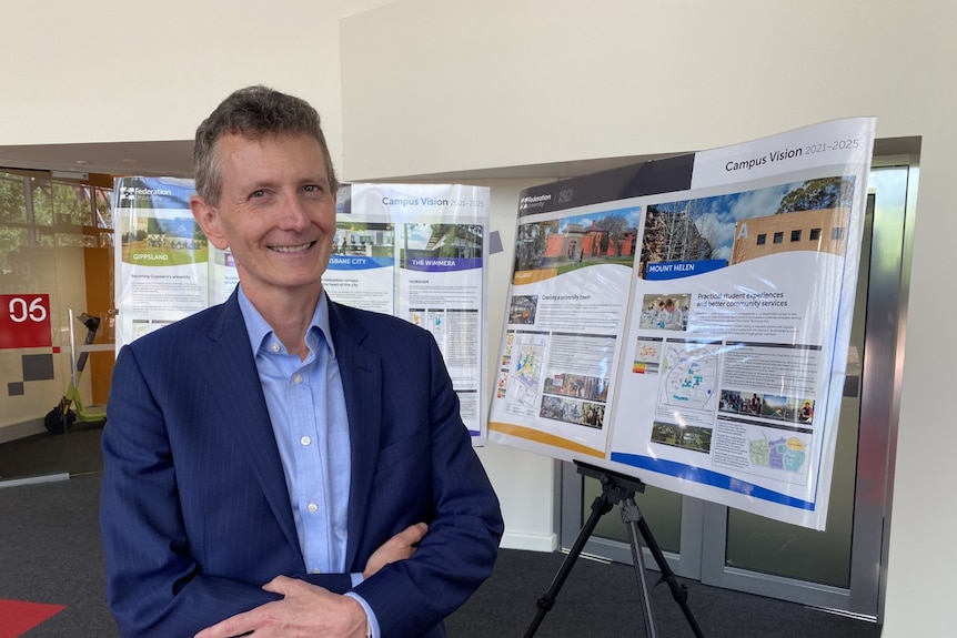 a man in a suit stands in front of a board with big photos and information about the new campus vision.
