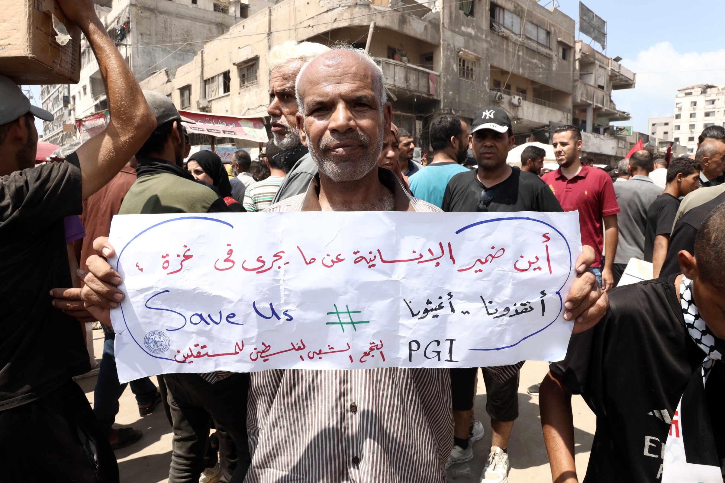 A man standing in a crowd, holding a sign written in Arabic with English words "Save Us".