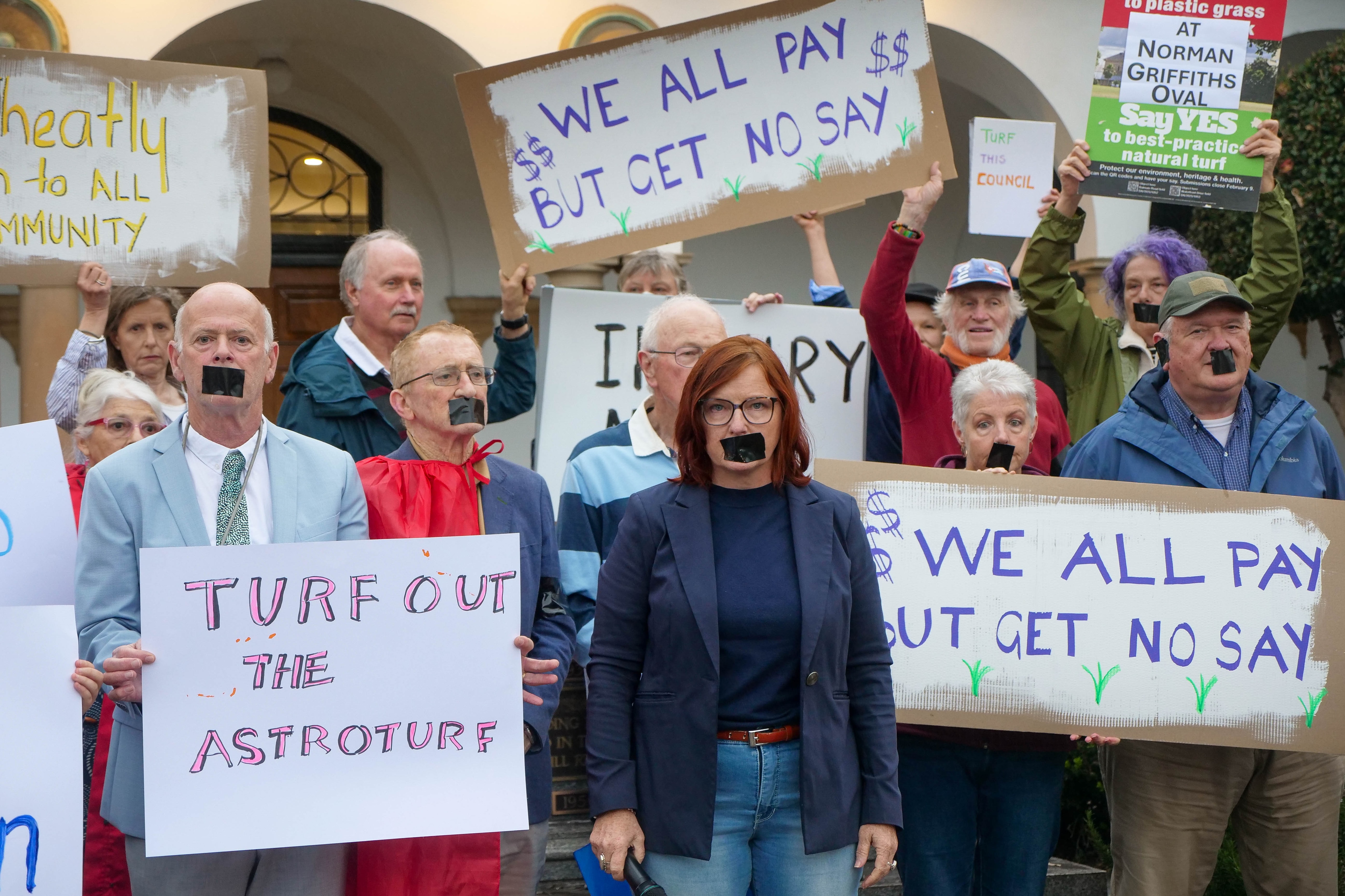 a small crowd holding signs against a synthetic football pitch, with tape over their mouths