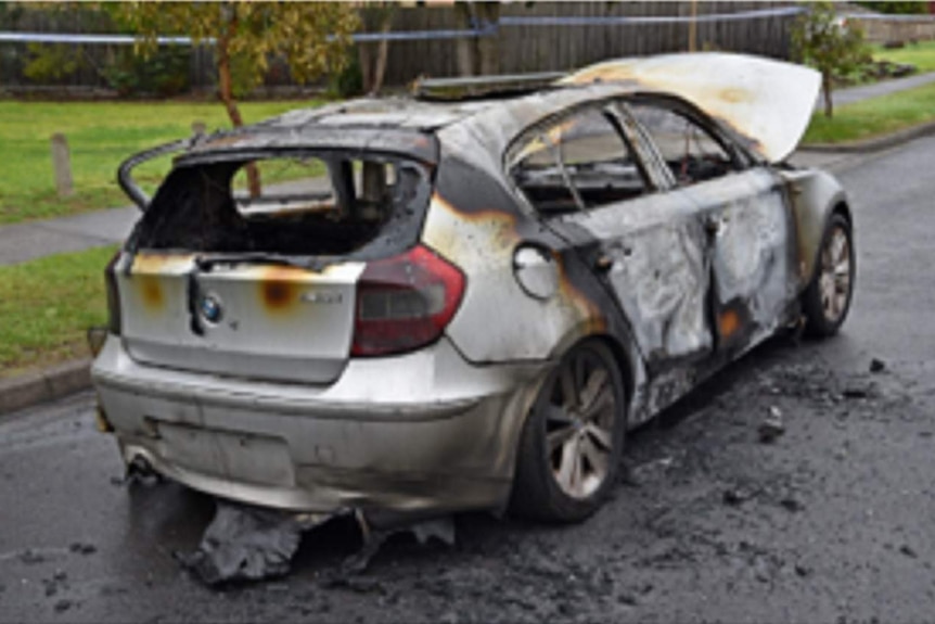 Police tape surrounds a charred and badly burnt BMW hatchback parked in a residential street.