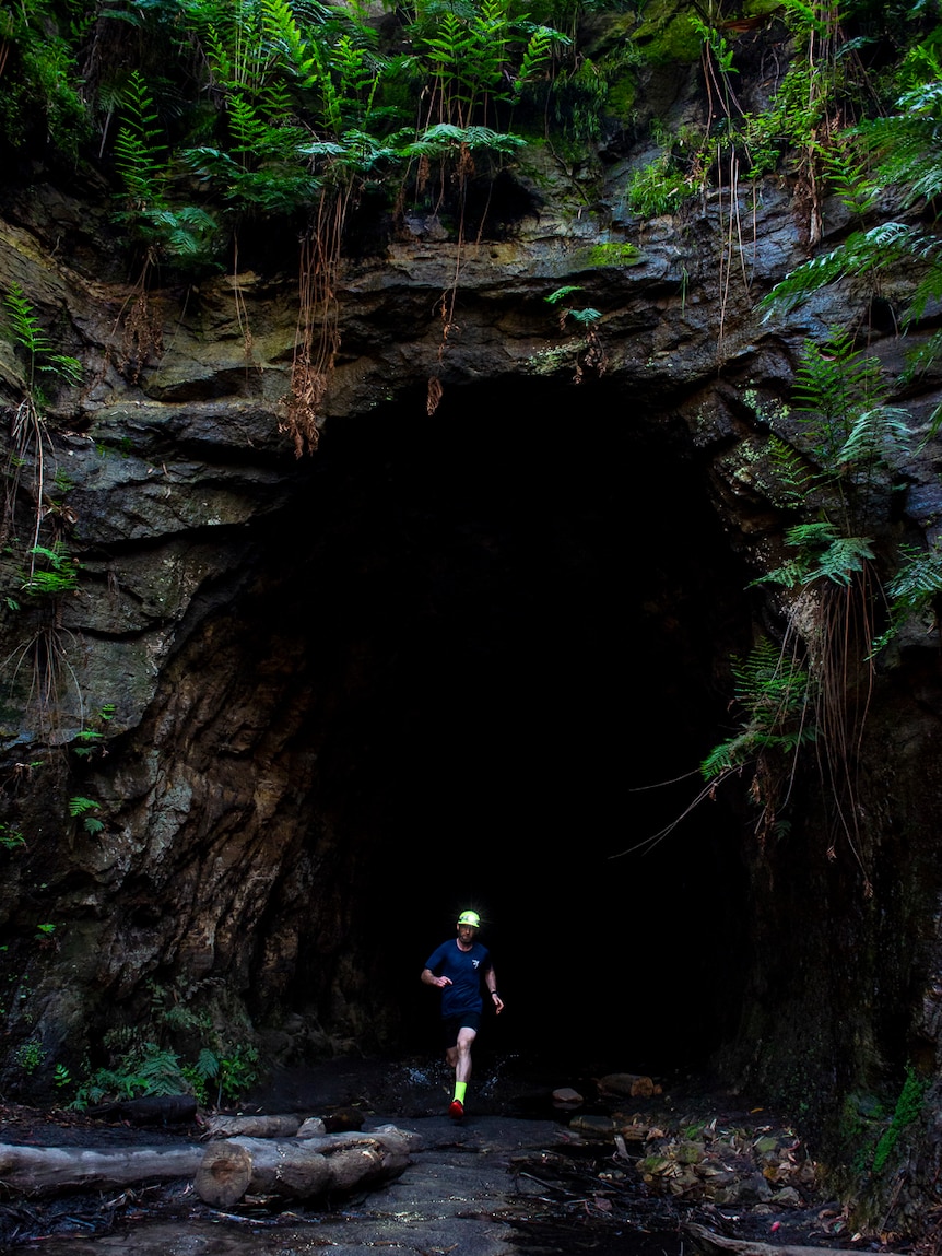 man with fluorescent hat and socks emerging from rocky tunnel covered in ferns