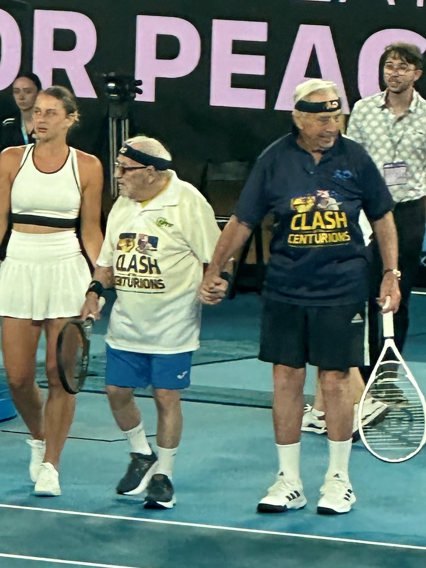 Two elderly male tennis players hold hands on court with racquets ready to play
