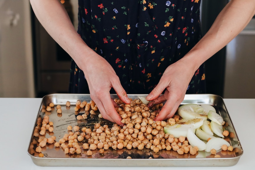 A pair of hands tosses chickpeas on a tray with sliced onion, oil and spices.