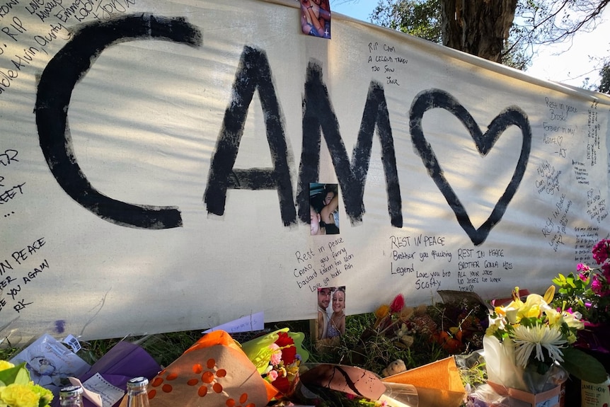 A white sheet reads CAM with a heart, and is covered with tribute messages and dozens of floral tributes beneath it.