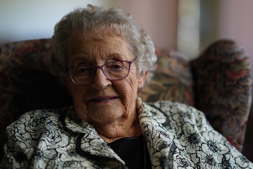 An elderly woman in purple glasses sits on a floral couch