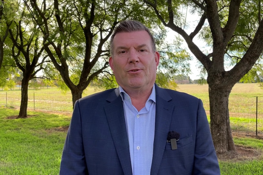  A man in a blue suit and shirt stands outdoors and talks. 