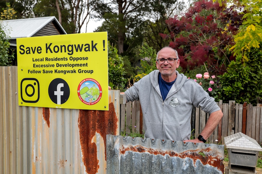 Man with glasses and grey hooded jumper stands next to yellow sign reading Save Kongwak, affixed to corrugated iron fence
