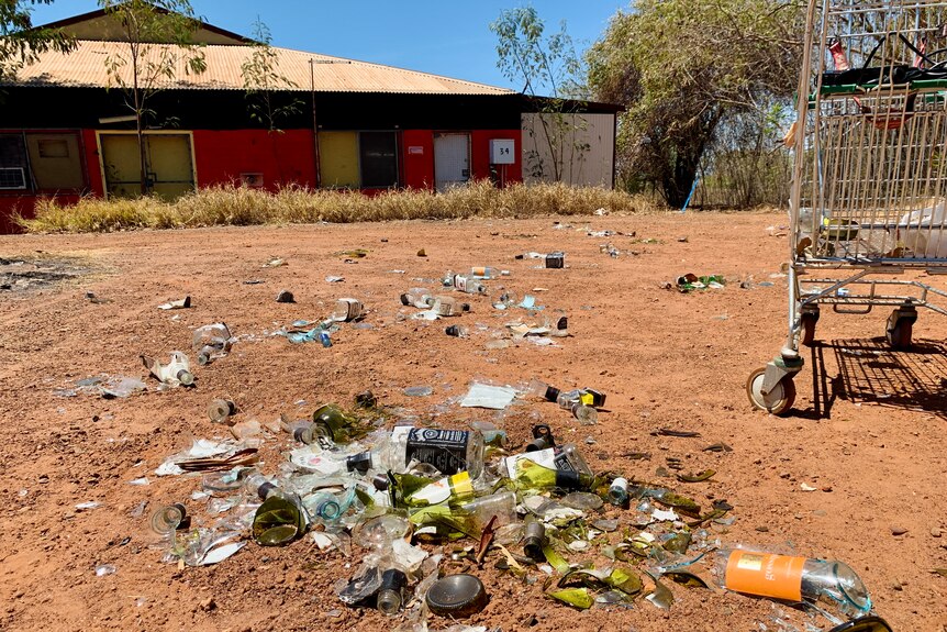 A pile of broken bottles and beer cartons lies in the red dirt in front of a house