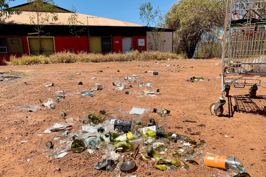 A pile of broken bottles and cartons of beer lies on the red earth in front of a house