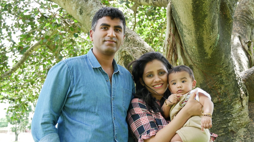 A young family of three stand outside, underneath a tree.